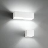 Two Vividara Wall Lights in white, embodying minimalist lighting and modern design, are mounted on a gray wall. They cast soft white light both upwards and downwards. The larger fixture positioned above the smaller one creates intersecting beams that form triangular patterns on the wall.