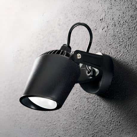 The Otis-1 Outdoor Single Spotlight in black is mounted on a textured grey wall. This durable lighting design includes a directional diffuser, casting light slightly downward, with visible wiring connecting to the bracket. Its sleek, minimalist style makes it ideal for both indoor and outdoor settings.