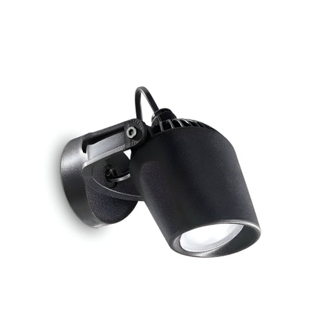 The Otis-1 Outdoor Single Spotlight in black is a contemporary fixture with a cylindrical design, angled downward. It boasts a sleek, industrial appearance, complete with a visible power cord and sturdy mounting bracket for reliable illumination. The light is currently switched off.