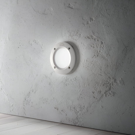 The Polar Recessed Round Outdoor Light - White, made from high-performance resin, is installed on a textured white wall. It provides soft illumination and is fastened with four visible screws around its edges. Beneath it lies a light-colored wooden floor, making it perfect for both indoor and outdoor lighting solutions.