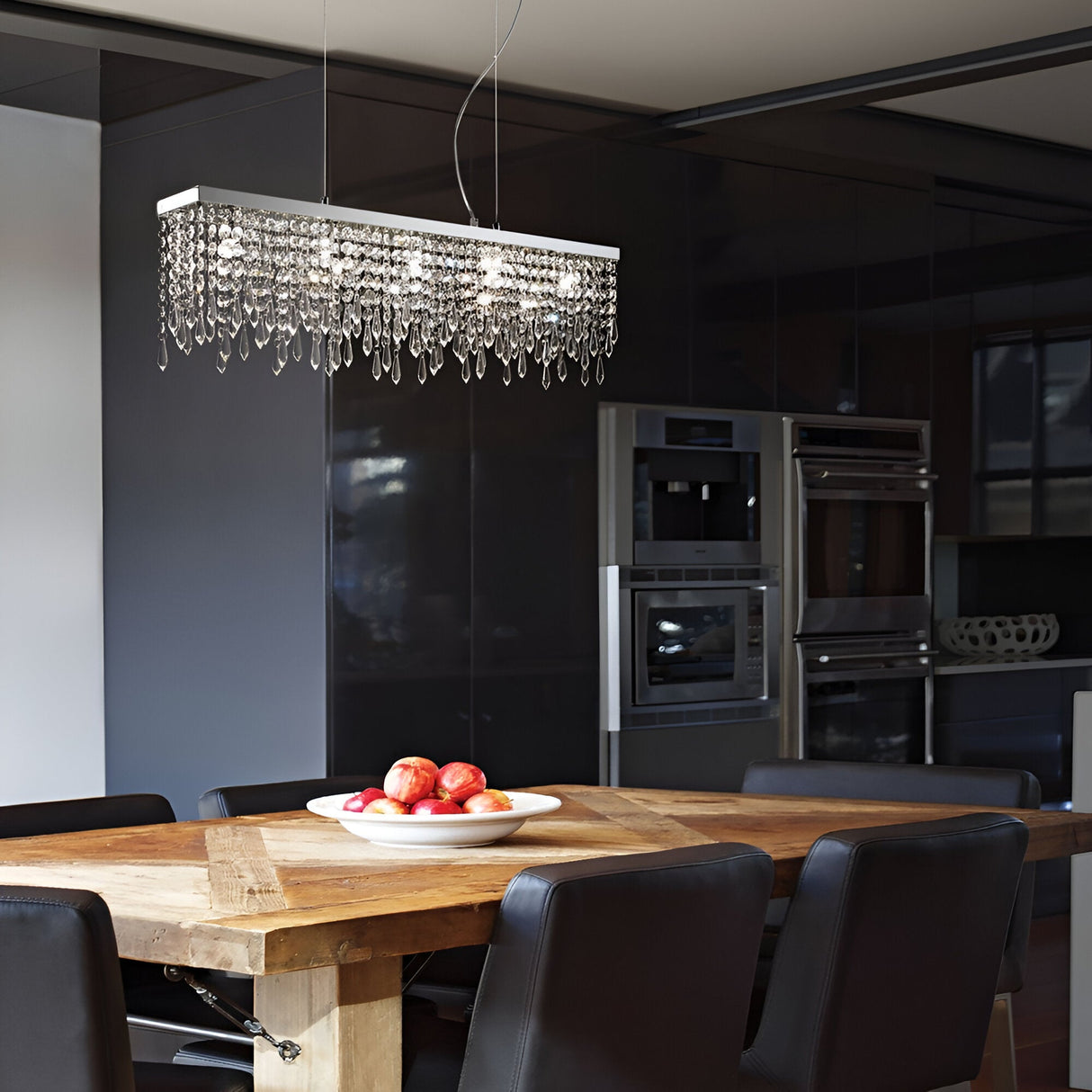 A modern dining room showcases a wooden table paired with black chairs. Elegance is added by the Iris 7 Light Crystal Suspension in Chrome hanging above, while a white plate filled with red apples sits on the table. The backdrop features sleek kitchen cabinetry and appliances accented in chrome.