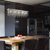 A modern dining room showcases a wooden table paired with black chairs. Elegance is added by the Iris 7 Light Crystal Suspension in Chrome hanging above, while a white plate filled with red apples sits on the table. The backdrop features sleek kitchen cabinetry and appliances accented in chrome.