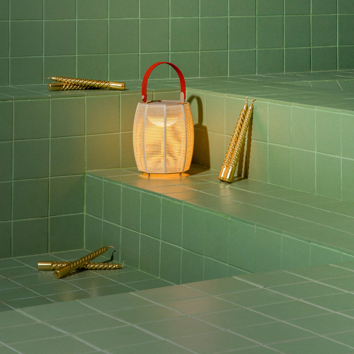 Tanit M/29/R rechargeable table lamp with woven beige fabric shade, red carry handle, and slim feet, lit on green tiled steps beside gold spiral candles.