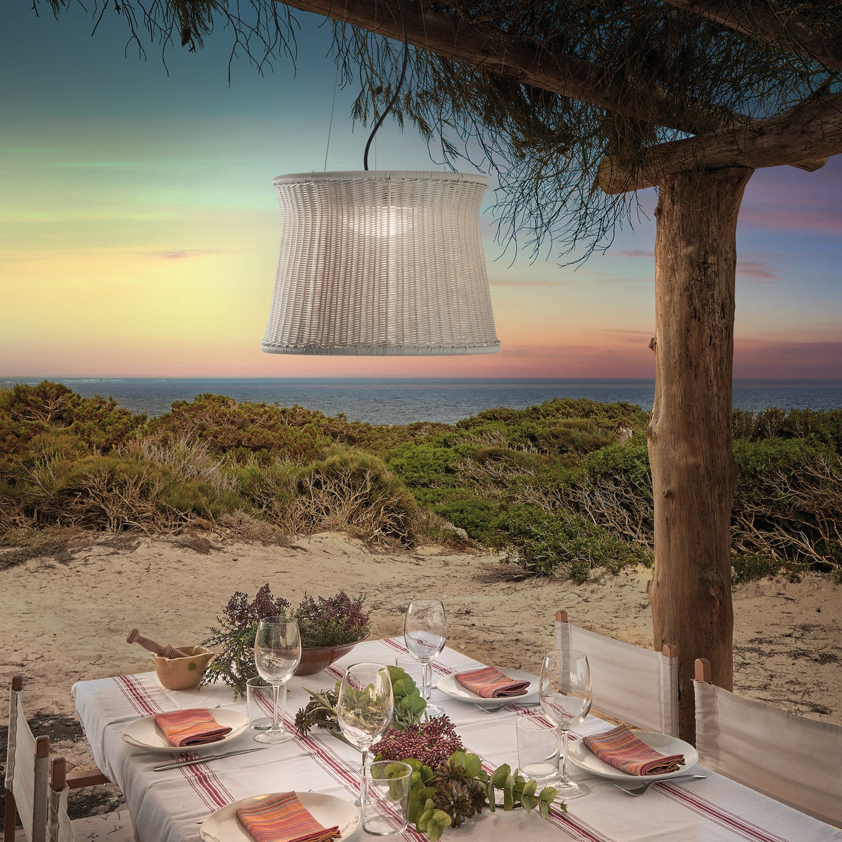 Syra 60 Aluminium LED Outdoor Pendant Light with woven white drum shade hanging over a coastal dining table at dusk, tree-suspended by fine wires.