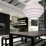 Mos 02 Ribbon Tiered Pendant Light with white pleated tiered shade on an iron frame, hung above a dining table in a modern kitchen and living space.