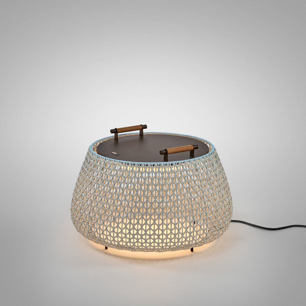 Nans Bag/38 Wood LED outdoor floor lamp with a wide, rounded woven beige shade, flat round dark top, and two teak carry handles, glowing warmly on a plain light grey background.