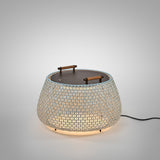 Nans Bag/38 Wood LED outdoor floor lamp with a wide, rounded woven beige shade, flat round dark top, and two teak carry handles, glowing warmly on a plain light grey background.