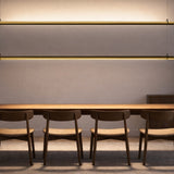 Solid SH 100 Tube LED Pendant Light in brass, two slim horizontal tubes suspended on fine wires above a wooden dining table with chairs, warm white glow against a light grey wall.