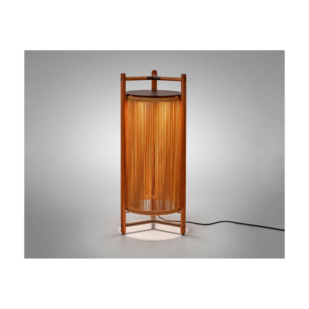 Kando P/111 outdoor floor lamp, lantern-style cylinder of sand nautical cord within a teak wood frame, topped with a dark brown disc, glowing warmly on a plain light grey background.