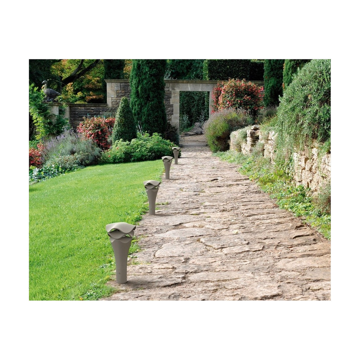Cornet B/52 Cone LED Bollard Light in stone grey lining a stone garden path, with a slender flared cone base and wavy layered cap.