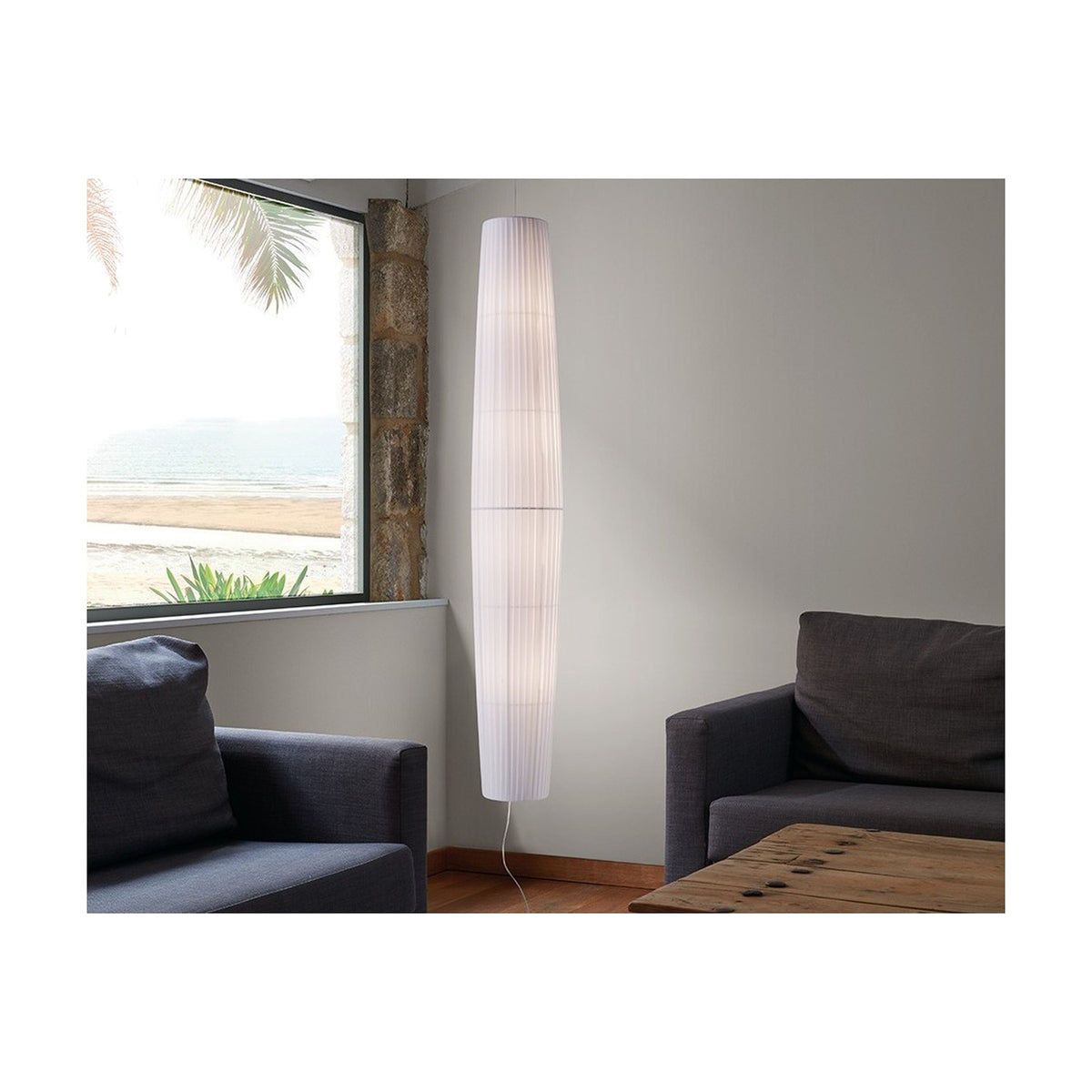 Maxi S/01 Ribbon LED pendant light with tall, tapered white pleated shade hanging beside a window in a living room with dark grey sofas.