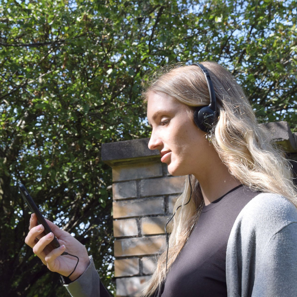 Woman outdoors wearing AV:Link Lightweight Stereo Headphones in black, wired to a phone in her hand, with trees and a brick pillar behind.