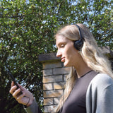Woman outdoors wearing AV:Link Lightweight Stereo Headphones in black, wired to a phone in her hand, with trees and a brick pillar behind.