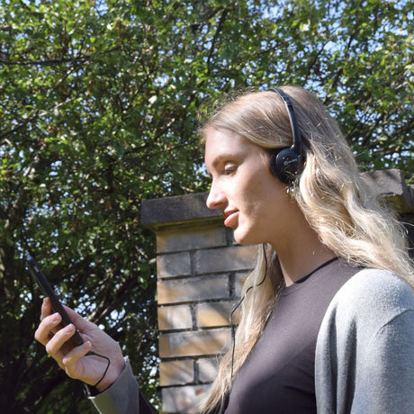Woman outdoors wearing AV:Link Lightweight Stereo Headphones in black, wired to a phone in her hand, with trees and a brick pillar behind.
