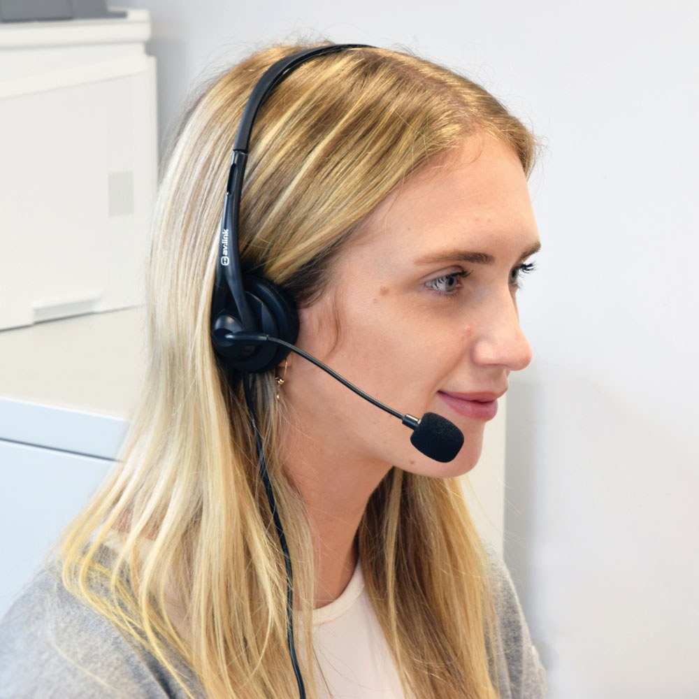 Blonde woman in an office wearing the AV:Link Multimedia Headset with black padded earcup and adjustable boom microphone positioned near her mouth.