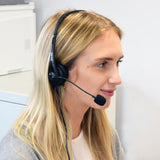 Blonde woman in an office wearing the AV:Link Multimedia Headset with black padded earcup and adjustable boom microphone positioned near her mouth.