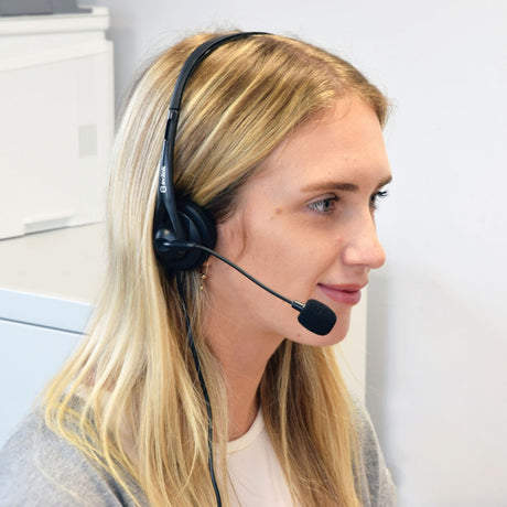 Blonde woman in an office wearing the AV:Link Multimedia Headset with black padded earcup and adjustable boom microphone positioned near her mouth.