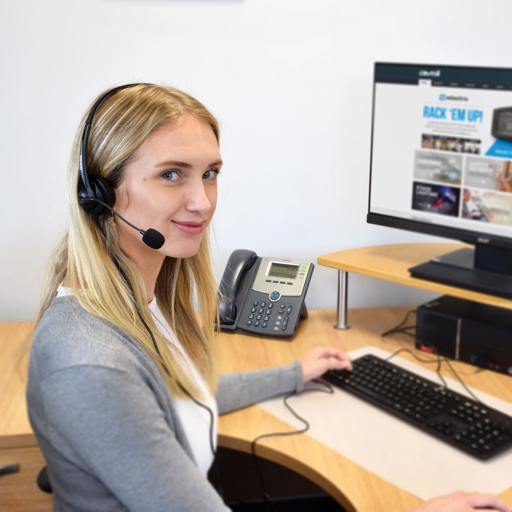 Blonde woman at a desk wearing the AVSL AV:Link multimedia headset with boom microphone, using a keyboard beside a desk phone and monitor.