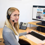Blonde woman at a desk wearing the AVSL AV:Link multimedia headset with boom microphone, using a keyboard beside a desk phone and monitor.
