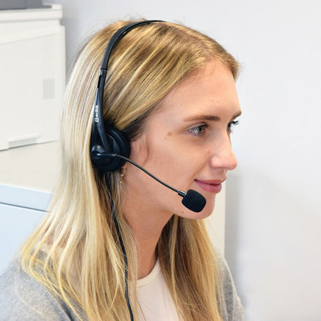 Woman in an office wearing the AV:Link USB Multimedia Headset, a black on-ear headphone with padded earcup and omnidirectional boom microphone.