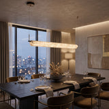 Anellara LED linear alabaster chandelier, 100cm horizontal bar with continuous veining, black canopy and dual cable suspension, shown above a dining table at dusk.