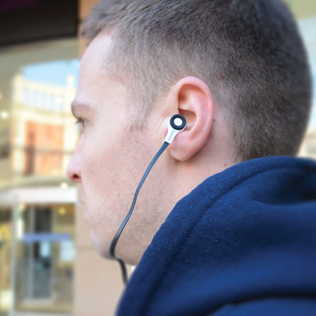 Side profile of a man wearing an AV:Link Rubberised Stereo Earphone with white housing and black flat cable in his ear outdoors.
