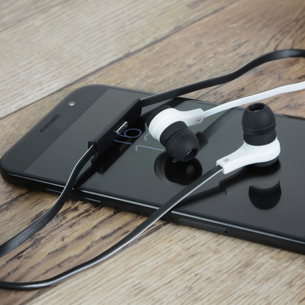AV:Link rubberised stereo earphones with white and black tips, flat black cable and inline mic, resting on a black smartphone on a wooden surface.