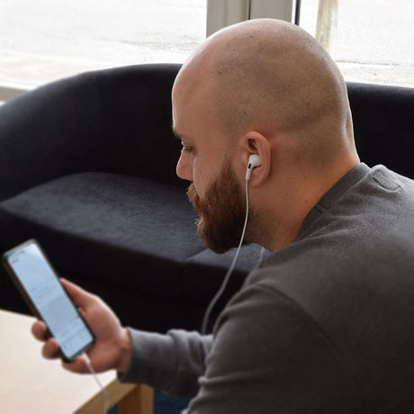 Bald bearded man in a grey top uses AV:Link USB Type-C wired in-ear earphones by AVSL, white earbud in ear with cable running to a phone.