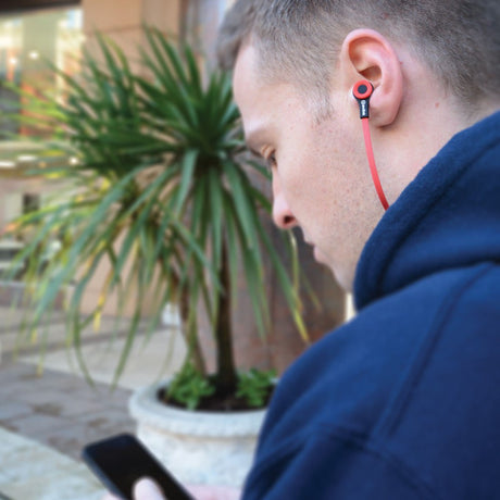 Man in navy hoodie wearing AV:Link Rubberised USB-C Stereo Earphones in red with flat cable, using a phone outdoors beside a potted palm.