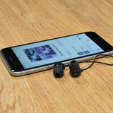 AV:Link EM9B black round mini in-ear earphones with 3.5mm cable resting beside a smartphone displaying a music app on a wooden surface.