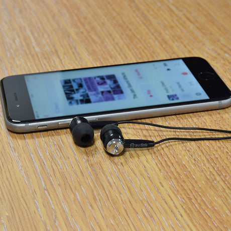 AV:Link EC9B black in-ear stereo earphones with silicone tips and 3.5mm jack lead, resting on a wooden desk beside a smartphone showing a music app.