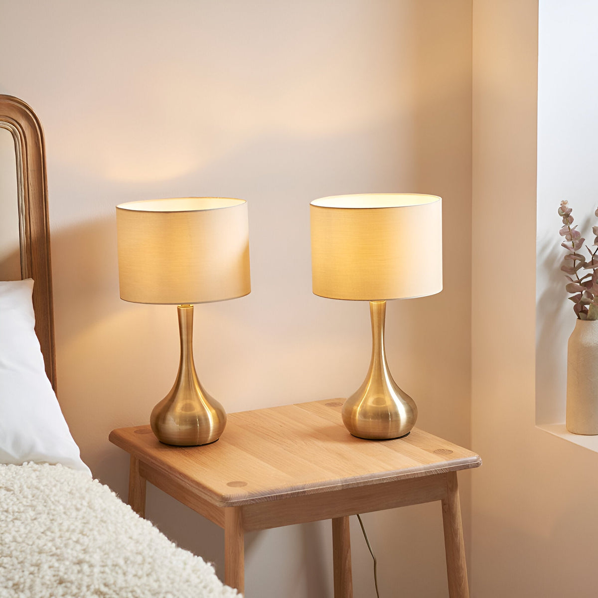 Endon Chartbury LED table lamps in brass with beige drum shades, shown as a twin pack on a wooden bedside table in a bedroom setting.