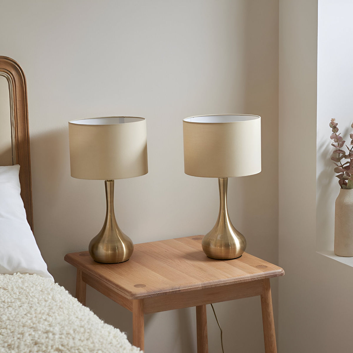 Endon Chartbury LED table lamps in brass with beige drum shades, shown as a pair on a wooden bedside table in a neutral bedroom.