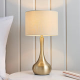 Endon Chartbury LED table lamp with brass teardrop base and beige drum shade on a bedside table beside a white pillow.