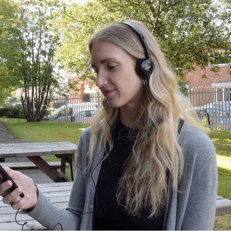 Woman wearing AV:Link SH30 lightweight stereo headphones outdoors, holding a phone connected by the 3.5mm cable, with trees and a bench behind.