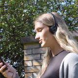 Woman outdoors wearing AV:Link SH30 lightweight fold-flat stereo headphones in black, with the wired 3.5mm cable running down to a phone in her hand.