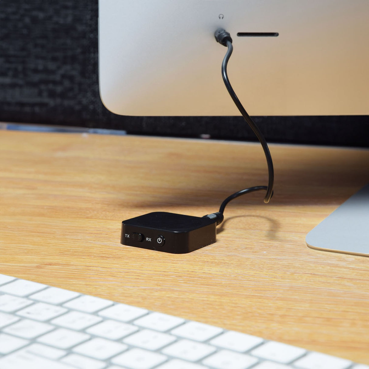 AV:Link BTTR2 Bluetooth audio transmitter and receiver in black, connected behind an iMac on a wooden desk with keyboard, TX/RX switch visible.
