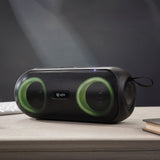 QTX PartyPod portable Bluetooth speaker with black mesh grille, twin drivers glowing green from the LED light show, and carry strap, shown on a wooden desk.