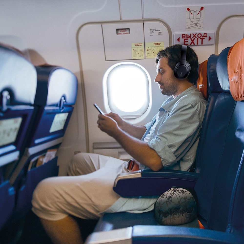 Man in aeroplane window seat wearing AV:Link Isolate SE black over-ear noise cancelling Bluetooth headphones while looking at his phone.