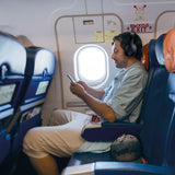 Man in aeroplane window seat wearing AV:Link Isolate SE black over-ear noise cancelling Bluetooth headphones while looking at his phone.