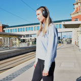 Woman at a train station platform wearing AVSL AV:Link Isolate SE black over-ear noise cancelling Bluetooth headphones with a light blue sweatshirt.