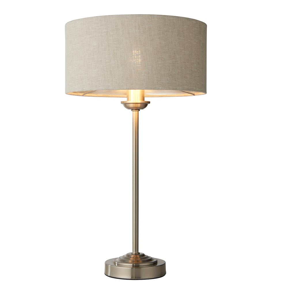 Brenley Table Lamp in brushed chrome with a natural linen drum shade on a slim metal stem and round base, shown lit against a white background.