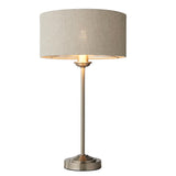 Brenley Table Lamp in brushed chrome with a natural linen drum shade on a slim metal stem and round base, shown lit against a white background.