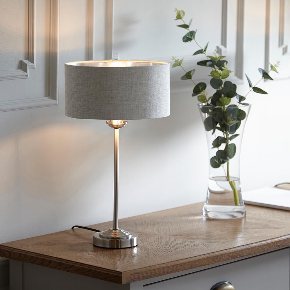 Brenley Table Lamp in brushed chrome with a grey fabric drum shade, shown lit on a wooden sideboard beside a glass vase with eucalyptus stems.