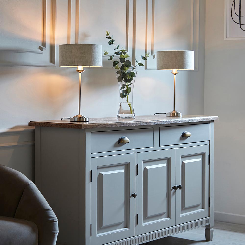 Pair of Brenley table lamps in brushed chrome with natural linen drum shades, styled on a grey sideboard beside a glass vase in a living room.