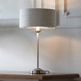 Brenley Table Lamp in brushed chrome with a grey linen drum shade, lit on a wooden console table against a panelled white wall.