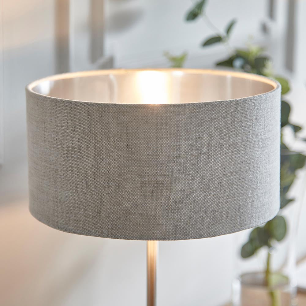 Brenley Table Lamp in brushed chrome with a grey linen drum shade and metallic inner lining, shown lit on a slim metal stem indoors.