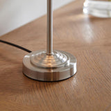 Close-up of the Brenley Table Lamp's brushed chrome base and stem on a wooden surface, showing the tiered circular foot and cable.