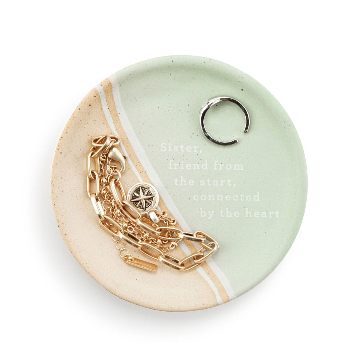 Round ceramic trinket dish in mint green and speckled beige with diagonal white stripe, "Sister, friend from the start" text, holding a ring and gold chain bracelet.