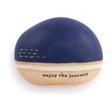 Smooth pebble-shaped ceramic ornament with a navy blue top, sand-coloured speckled base, white stripe, and "enjoy the journey" text, on a plain white background.